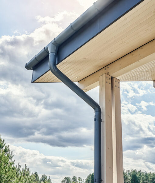 Contemporary grey metal rainwater downpipe installed on roof of new building with wooden terrace on cloudy day close view